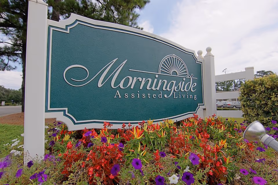 A green sign that reads "Morningside Assisted Living" mounted by a white post with colorful flowers in front and a fence behind it.