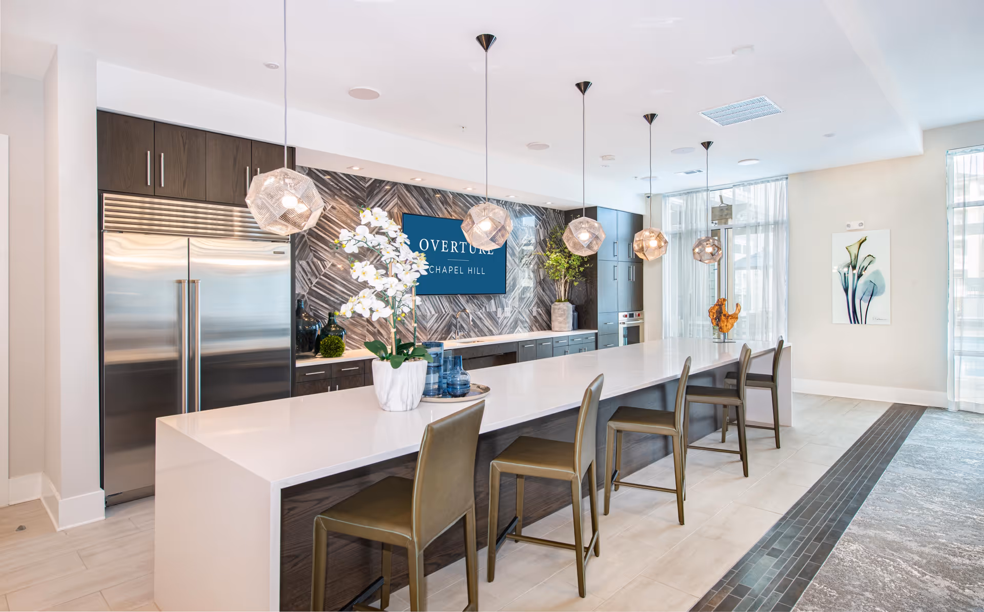 Bright modern community kitchen with a long white island, bar stools, pendant lights, stainless steel refrigerators and a sign reading "Overture Chapel Hill" on the back wall.