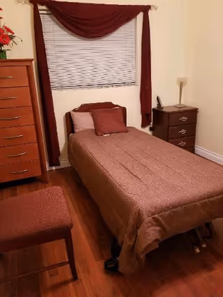 A small bedroom with a single bed covered in a brown bedspread and matching pillows. There is a wooden dresser on the left side, a nightstand with a lamp and phone on the right side, and a window with closed blinds and maroon curtains above the bed. A matching brown cushioned bench is partially visible in the foreground.