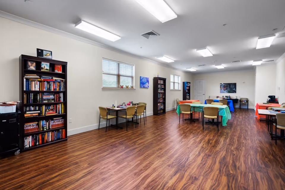 A spacious room with wood-patterned flooring and white walls, featuring several tables covered with colorful tablecloths and surrounded by chairs. There are bookshelves filled with books along the walls, windows letting in natural light, and fluorescent ceiling lights illuminating the room.