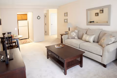 A bright living room with a beige sofa adorned with patterned cushions, a dark wooden coffee table with magazines on it, a side table with a lamp, and a large mirror on the wall above the sofa. In the background, there is a small dining area with a table and chairs, and a kitchen with wooden cabinets and a white refrigerator.
