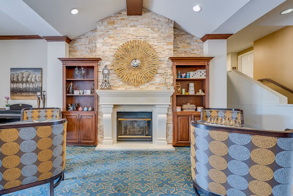 A cozy senior living common area with a stone fireplace centered between two wooden bookshelves filled with decorative items. Two patterned armchairs face the fireplace, and a chessboard is placed on one of the chairs. The room has a blue floral carpet and a sunburst wall decoration above the fireplace.