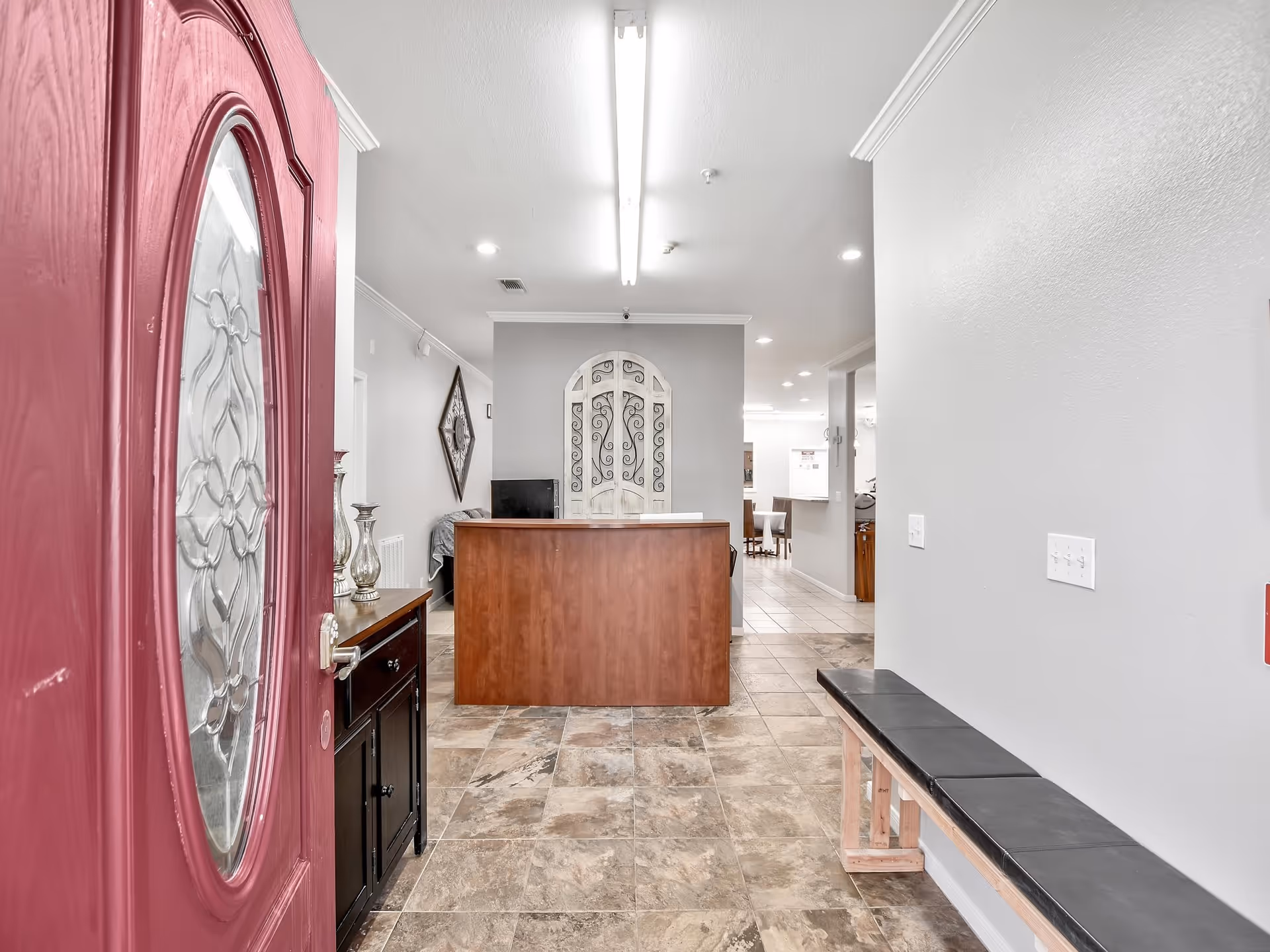 View into an assisted living facility reception area with an open red front door, tiled floor, wooden reception desk, and a bench along the wall.
