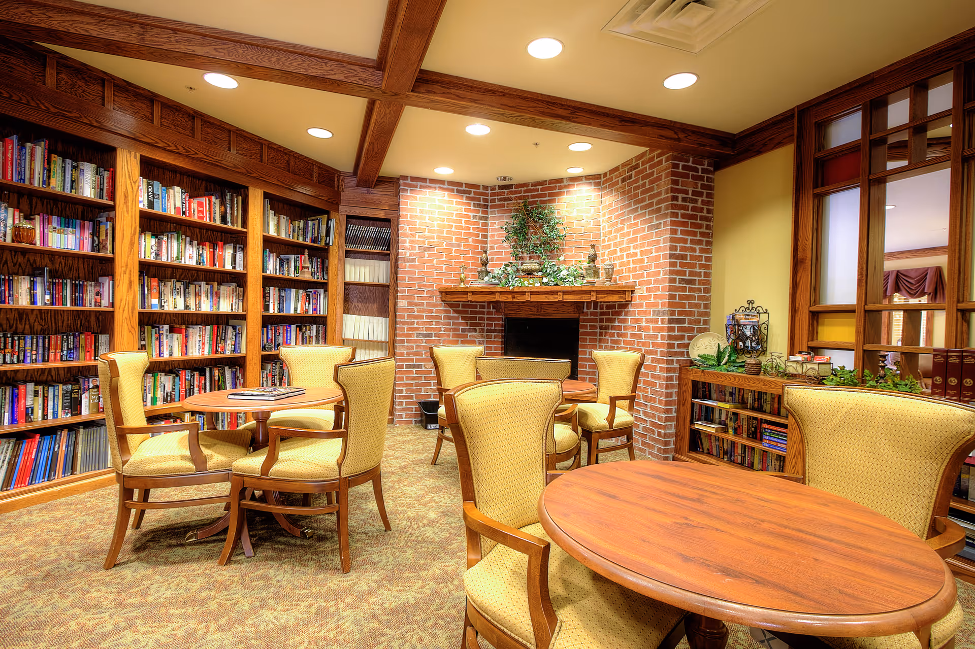 A cozy library room with wooden bookshelves filled with books, several round wooden tables surrounded by yellow upholstered chairs, a brick fireplace decorated with plants, and warm lighting from ceiling recessed lights.
