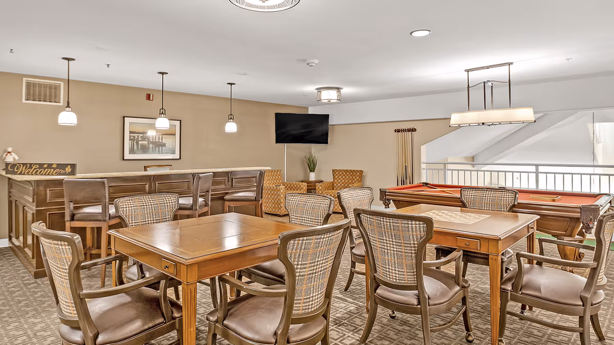 A well-lit recreational room with two wooden game tables surrounded by cushioned chairs, a pool table with a red felt surface, a bar area with high stools, two patterned armchairs, a wall-mounted TV, and a 'Welcome' sign on the bar counter.