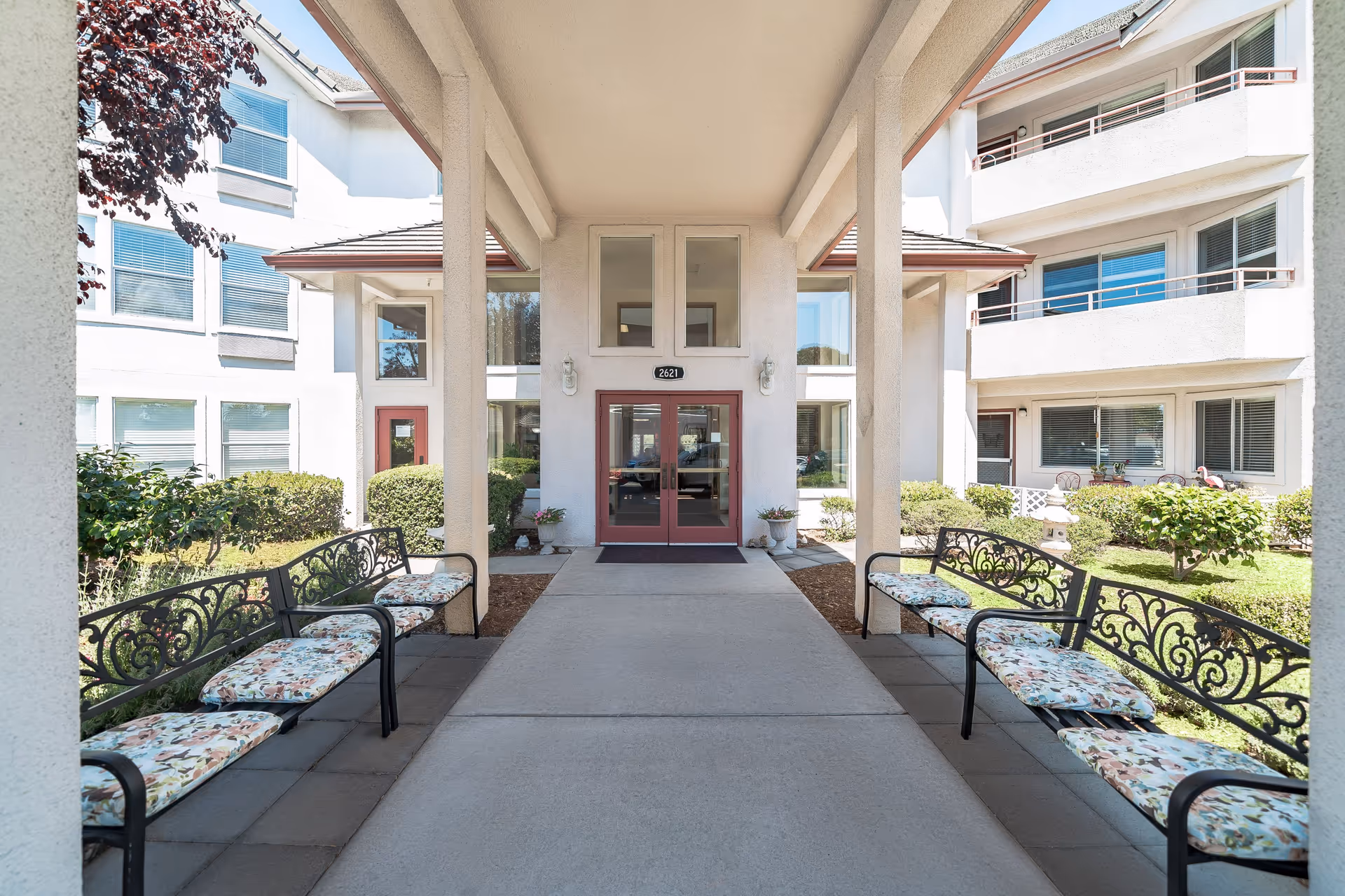 Covered entrance walkway leading to double glass doors of a senior living facility building. There are decorative black metal benches with floral cushions on both sides of the walkway. The building is white with multiple windows and some greenery around the entrance.