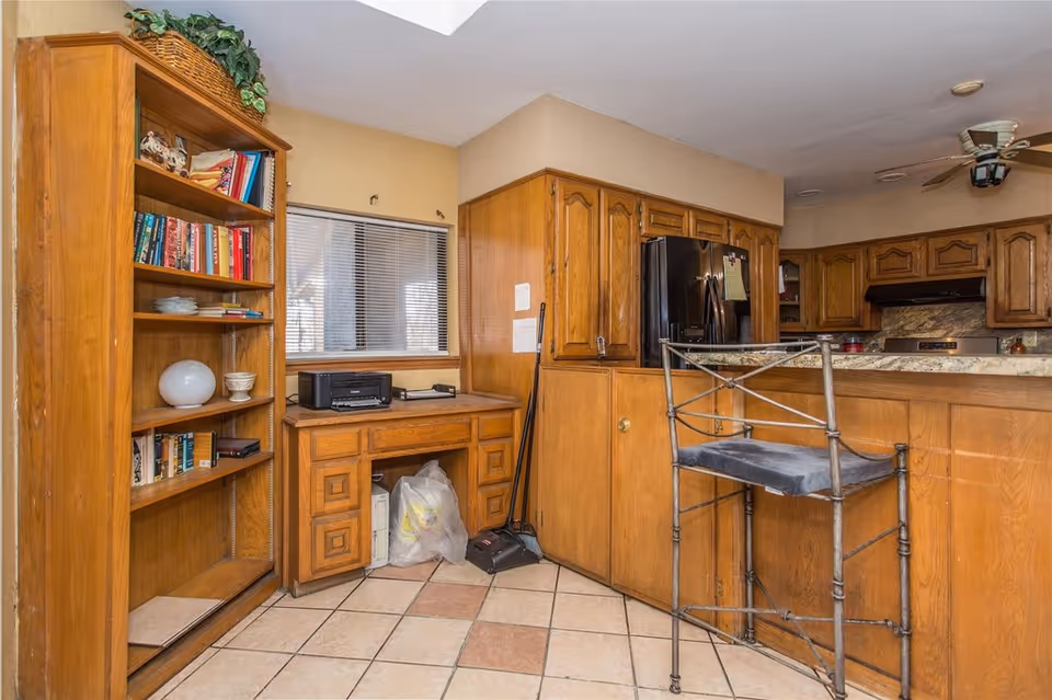 Wood-paneled kitchen area with wooden cabinets, a countertop bar with a metal stool, a bookshelf and a small desk on a tiled floor.