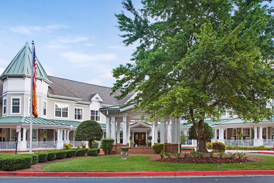Front entrance of Ivey Ridge Senior Living with a covered porte-cochère, flags, a large tree, and a wraparound porch on a multi-story building.