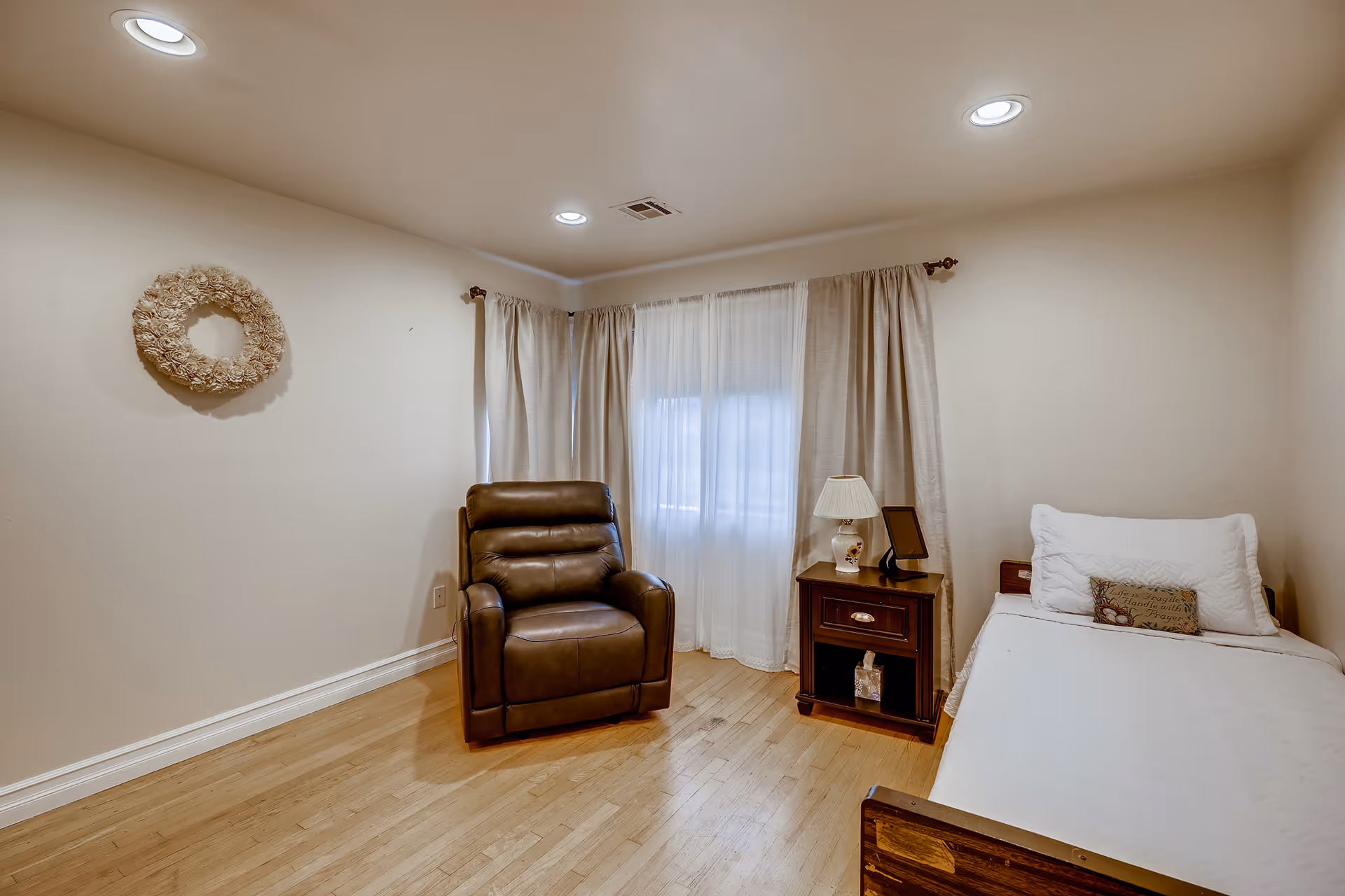 Cozy bedroom with a single bed, bedside table and lamp, a leather recliner, wood floors, and a curtained window.