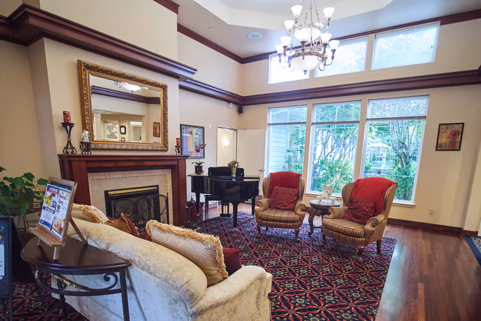 A cozy living room area with a patterned red carpet, two striped armchairs with red cushions, a small round wooden table between them, a beige sofa with multiple pillows, a grand piano near large windows letting in natural light, and a fireplace with a large ornate mirror above it.