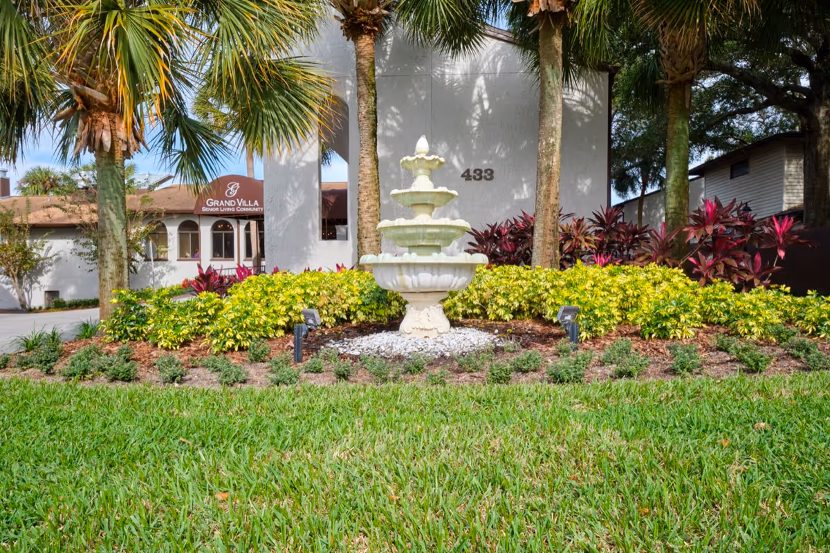 Exterior view of Grand Villa Senior Living Community building with a white three-tiered fountain in the foreground, surrounded by green grass, palm trees, and colorful shrubs. The building has the number 433 displayed on the wall.