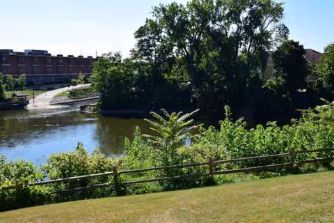 Grassy riverside with trees, a wooden fence, and distant buildings.