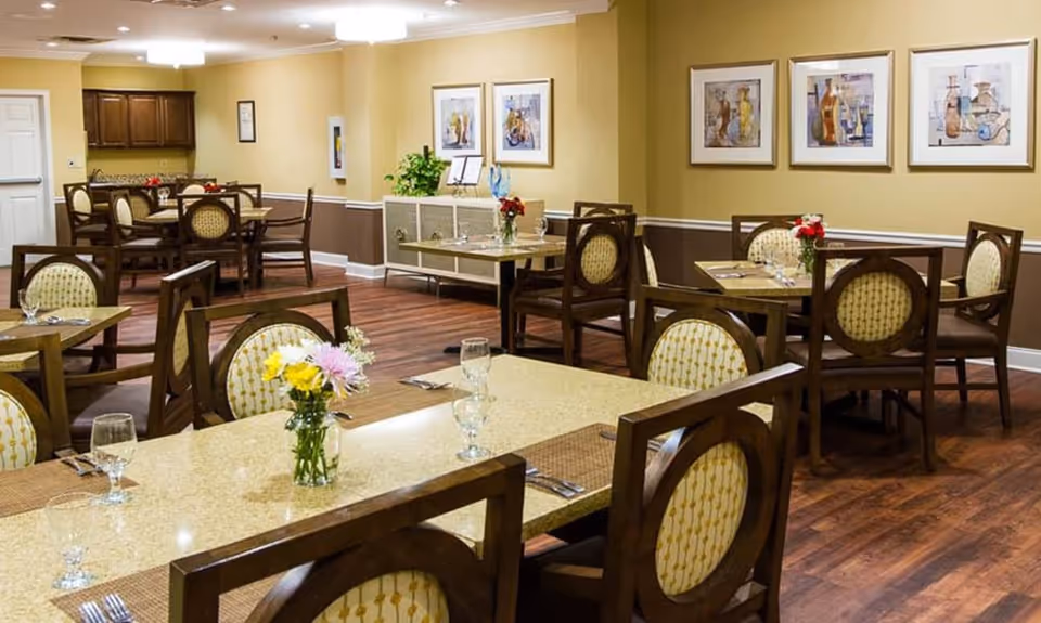 A dining room with multiple tables and chairs arranged neatly. Each table has a small vase with flowers and place settings including glasses and utensils. The room has warm yellow walls with framed artwork and wooden flooring.