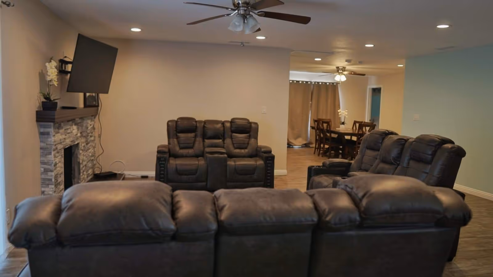 A cozy living room with dark leather recliner sofas arranged facing a wall-mounted flat screen TV above a stone fireplace. The room has ceiling fans with lights, recessed lighting, and wood flooring. In the background, there is a dining area with a wooden table and chairs, and beige curtains covering a window or door.