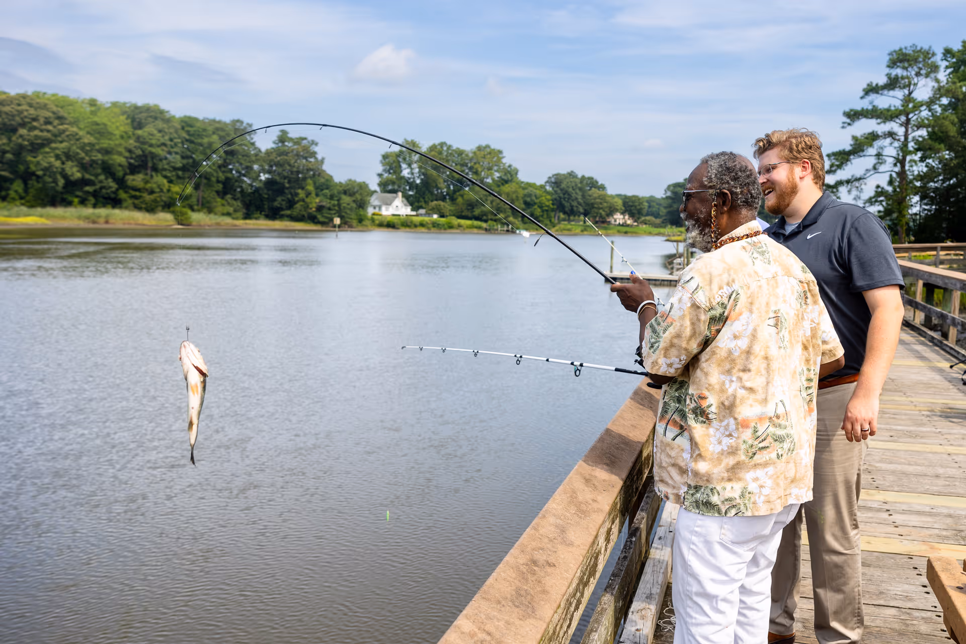 An elderly man and a younger man are fishing together on a wooden dock by a calm river. The elderly man is holding a fishing rod with a fish caught on the line, while the younger man stands beside him smiling. Trees and houses are visible in the background under a partly cloudy sky.