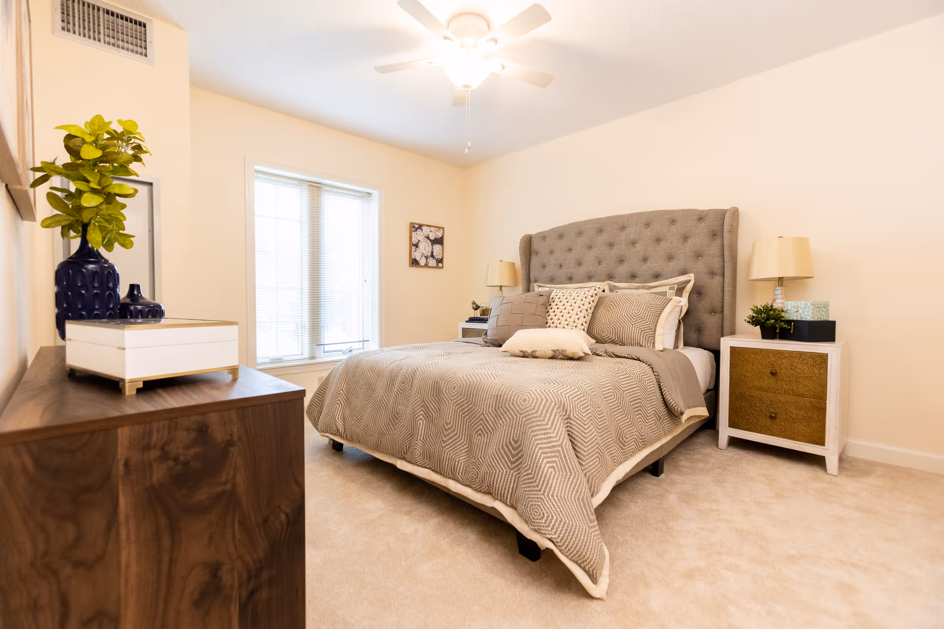 Bright, neatly furnished bedroom with a tufted headboard, bed, nightstands, dresser, and a window.