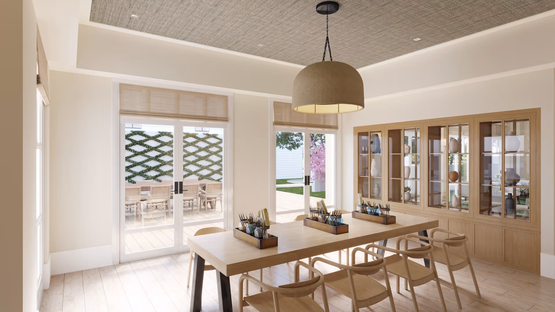 A bright and airy room with a long wooden table surrounded by six wooden chairs. On the table are containers holding paintbrushes and art supplies. Behind the table is a large wooden cabinet with glass doors displaying decorative vases. Large glass doors open to an outdoor patio area with a table and chairs, and greenery is visible outside. A large, round pendant light hangs from the ceiling.