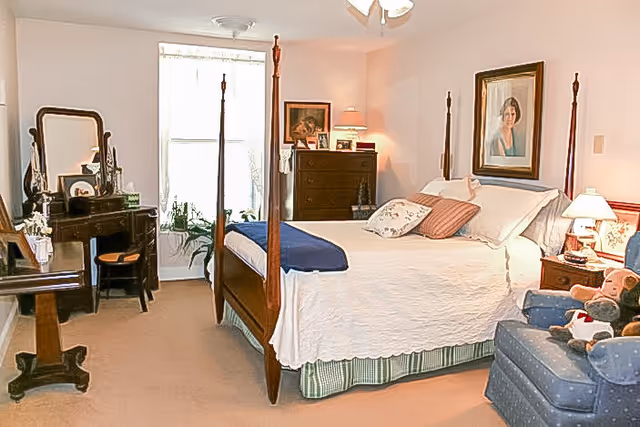 A cozy bedroom with a four-poster bed covered in white bedding and decorative pillows. There is a wooden dresser with framed photos and a lamp, a vanity with a mirror and a chair, and a blue upholstered armchair with stuffed animals. The room is softly lit with natural light coming through a window with sheer curtains.