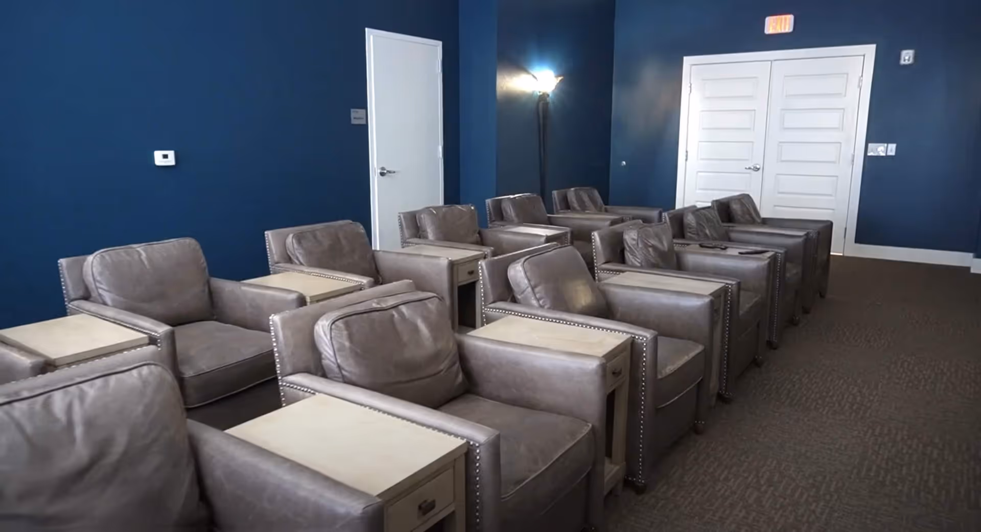 A room with two rows of gray leather armchairs separated by small wooden side tables. The walls are painted dark blue, and there are white doors and a floor lamp in the background.