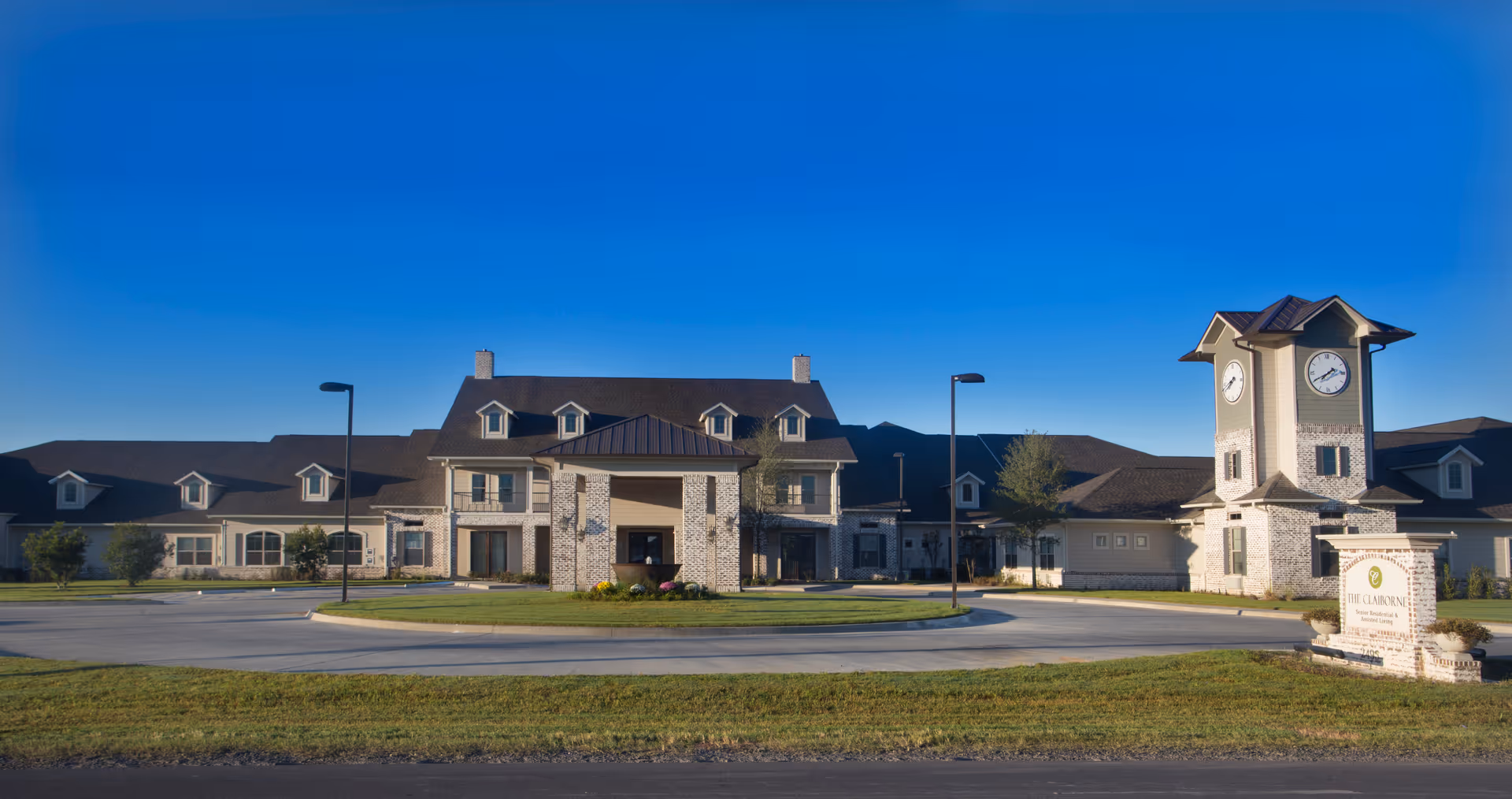 Front exterior view of The Claiborne at Thibodaux senior living facility with a large building featuring multiple windows, a covered entrance, and a clock tower on the right side. The sky is clear and blue, and there is a circular driveway with street lamps and some landscaping in front.