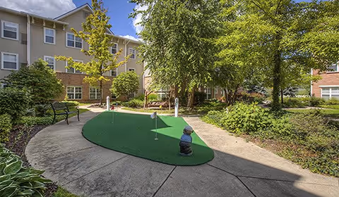 Outdoor courtyard area in a senior living facility with a putting green, benches, trees, and shrubs surrounded by a multi-story building under a partly cloudy sky.
