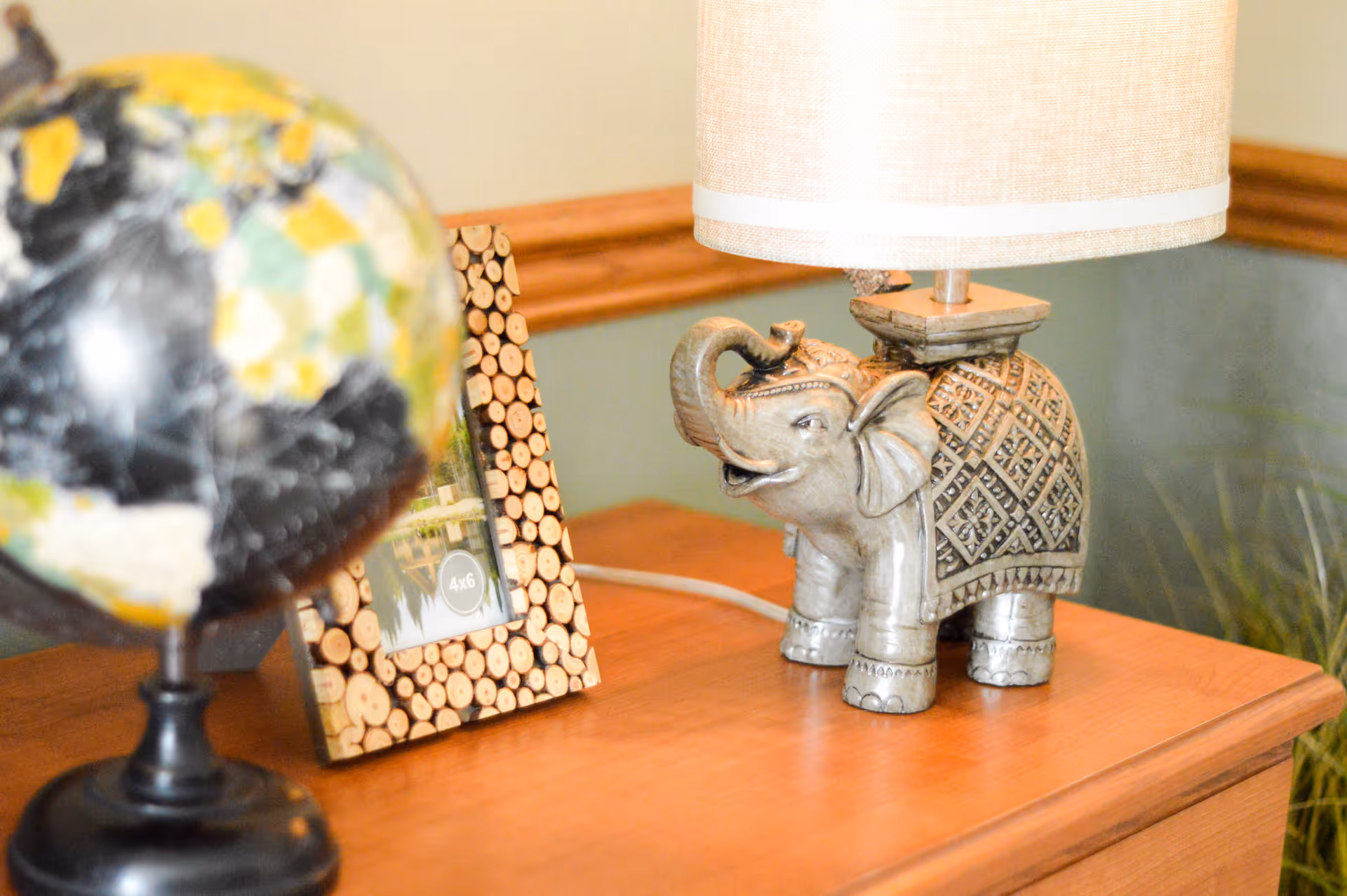 A wooden table with a decorative globe, a photo frame made of small wooden discs, and a lamp with a base shaped like an intricately carved elephant.