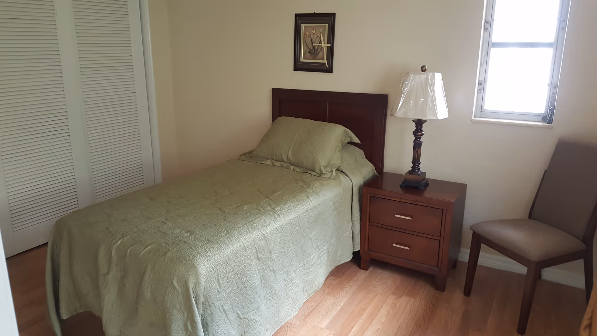 A small bedroom with a single bed covered in a green bedspread, a wooden headboard, a wooden nightstand with two drawers, a lamp with a white shade, a brown upholstered chair, a window, and a framed floral picture on the wall.