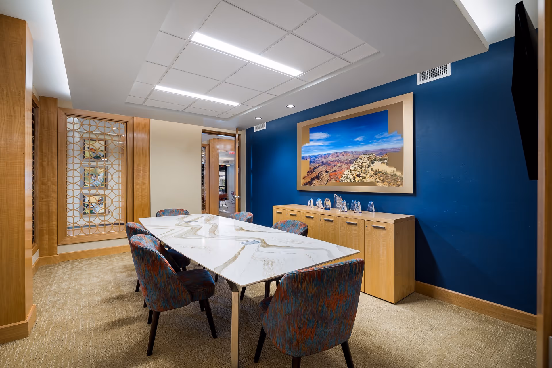 A modern conference or meeting room with a long white marble table surrounded by six upholstered chairs. The room features a blue accent wall with a large framed photograph of a canyon landscape above a wooden cabinet. The opposite wall has wooden paneling with decorative glass inserts. The ceiling has recessed lighting panels.
