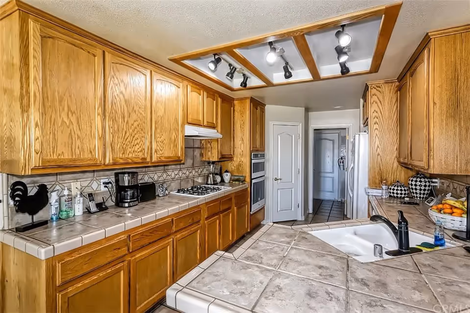 A kitchen with wooden cabinets, tiled countertops, a built-in gas stove, a coffee maker, a sink with a black faucet, and various kitchen items including a fruit basket and decorative containers. The ceiling has a recessed lighting fixture with multiple spotlights.
