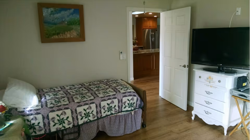 A small bedroom with a single bed covered in a patterned quilt. There is a white pillow on the bed and a framed painting hanging on the wall above it. A white dresser with floral designs holds a flat-screen TV. The door is open, revealing a kitchen area with wooden cabinets and a stainless steel refrigerator. The floor is wooden throughout.