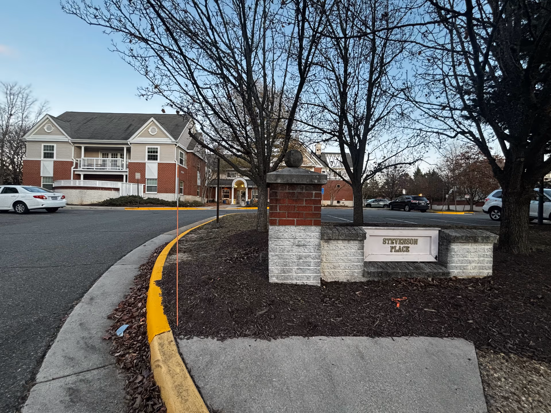 Exterior view of Stevenson Place senior living facility showing a brick and stone sign with the facility name, a curved driveway with yellow-painted curb, several parked cars, leafless trees, and a two-story building with red brick and beige siding under a clear sky.