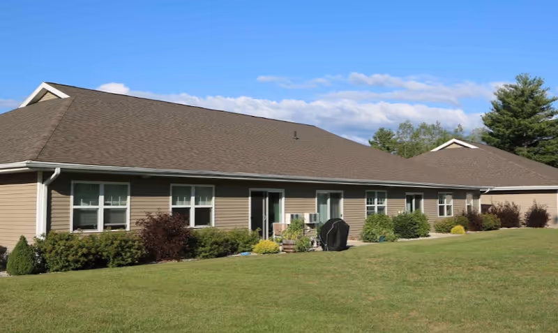 Single-story building with beige siding and a brown roof, surrounded by green grass and shrubs under a blue sky with some clouds.