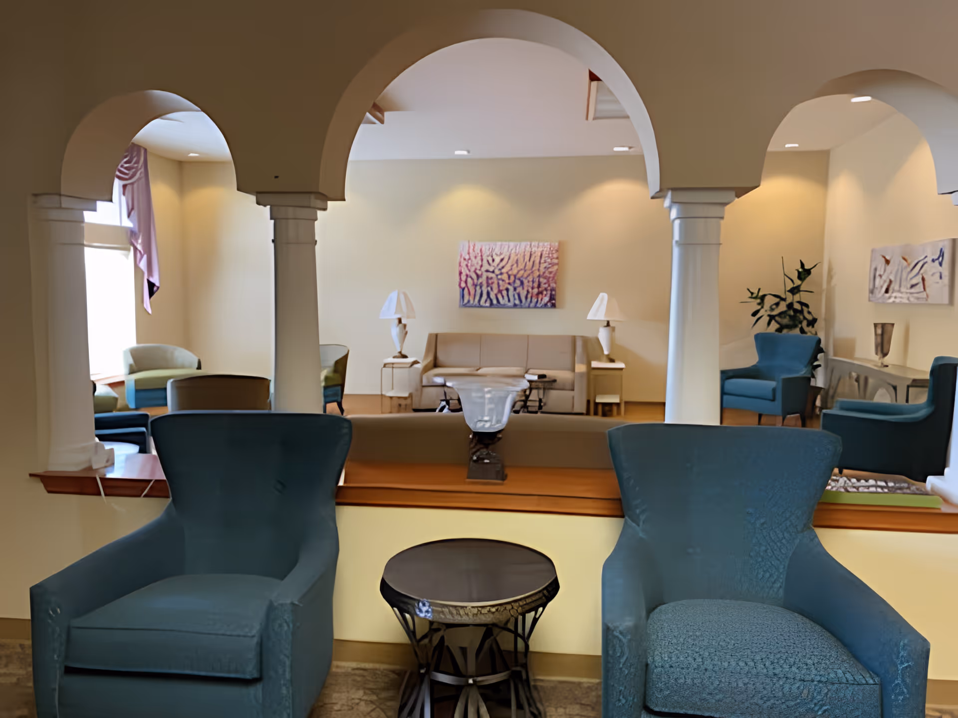 A cozy living room area with two blue armchairs in the foreground separated by a small round table. In the background, there is a beige sofa with two white lamps on side tables and colorful abstract paintings on the walls. The room features arched openings supported by white columns, and additional seating is visible through the arches.