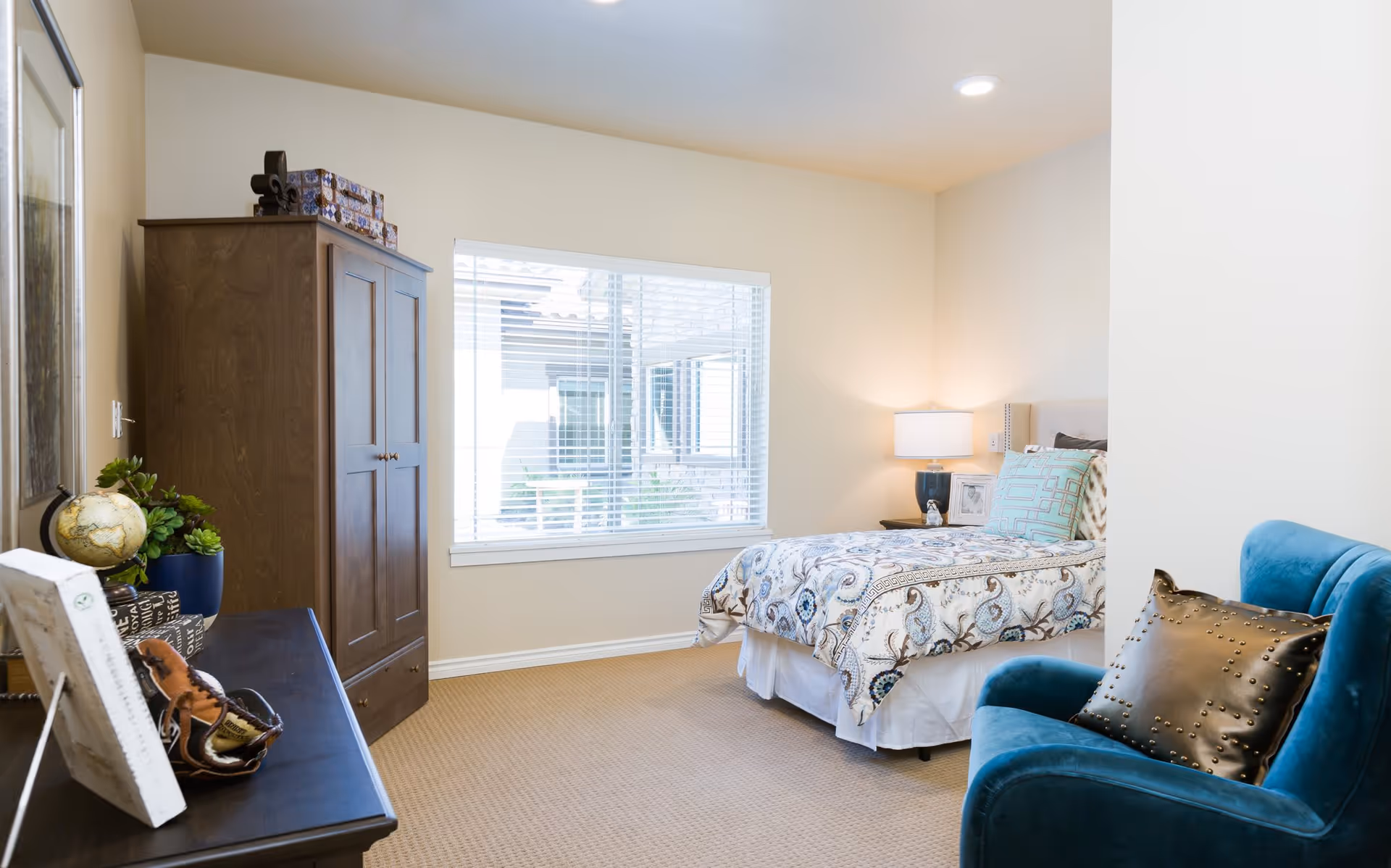 A bright bedroom with a bed covered in a patterned comforter, a wooden wardrobe, a blue armchair with a studded pillow, a nightstand with a lamp, and a window with white blinds letting in natural light.