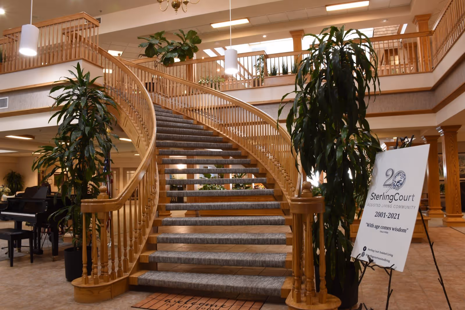 Interior view of an assisted living community featuring a wide, curved wooden staircase with carpeted steps. The area is decorated with large green plants, a black grand piano, and a sign celebrating 20 years of Sterling Court Assisted Living Community from 2001 to 2021. The space has warm lighting and wooden railings on the upper level.