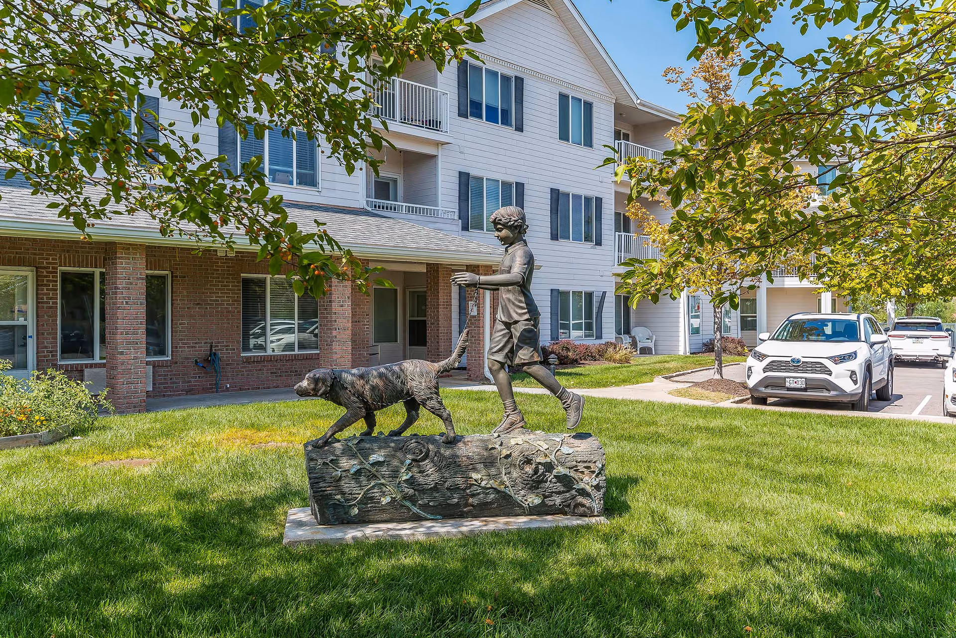 Outdoor scene at Solstice Senior Living at Columbia showing a bronze statue of a boy walking a dog on a log in a grassy area. The background features a multi-story residential building with balconies, windows, and parked cars along a driveway. Trees with green leaves frame the scene.