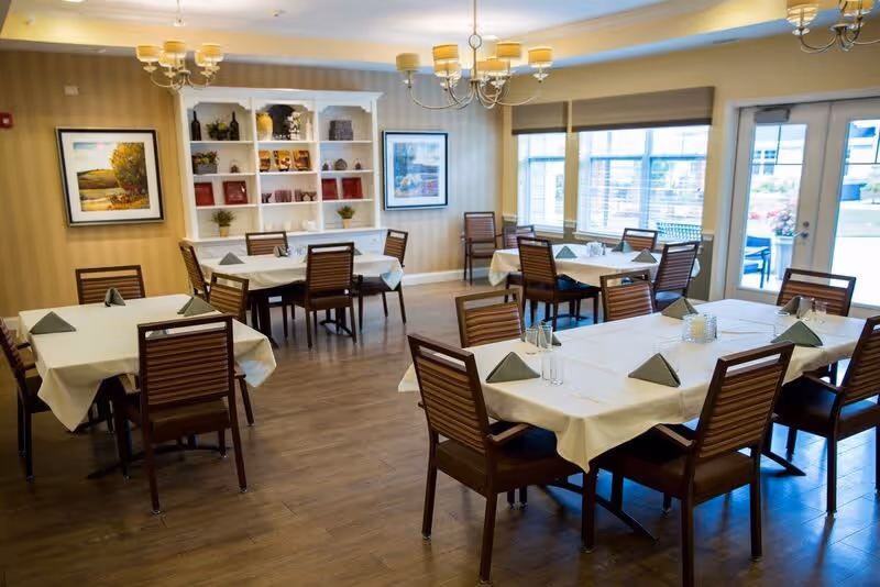 A bright and neatly arranged dining room with several tables covered in white tablecloths and set with folded green napkins and glassware. Wooden chairs surround the tables. The room features wood flooring, beige striped wallpaper, two chandeliers, framed artwork on the walls, a white shelving unit with decorative items, and large windows letting in natural light. A door leads to an outdoor area.