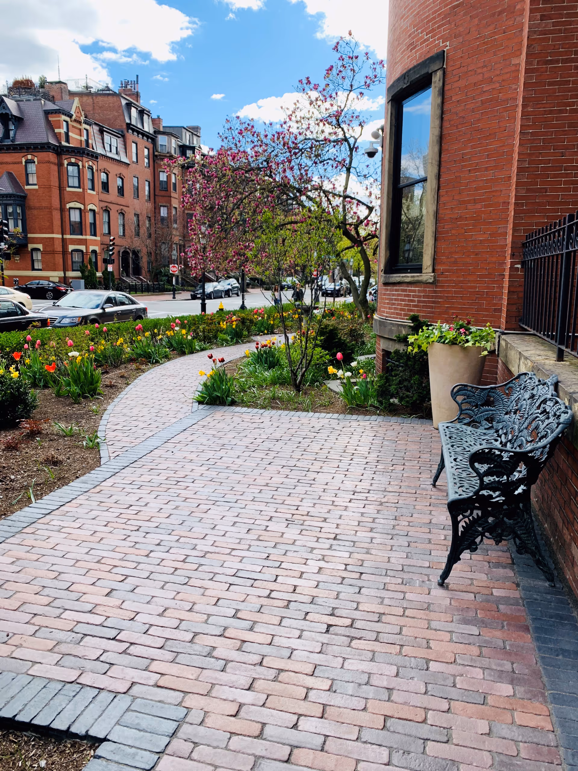 Brick-paved pathway and ornate metal bench beside a red-brick building with flowering tulips and a blooming tree facing a street of brownstone buildings.