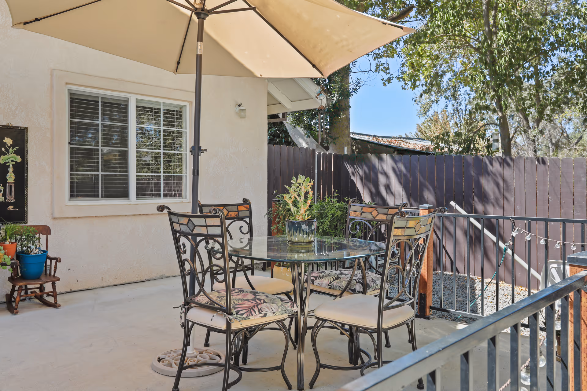 Outdoor patio with a glass-top dining table, four ornate metal chairs with cushions under a large umbrella, potted plants, and a wooden fence.