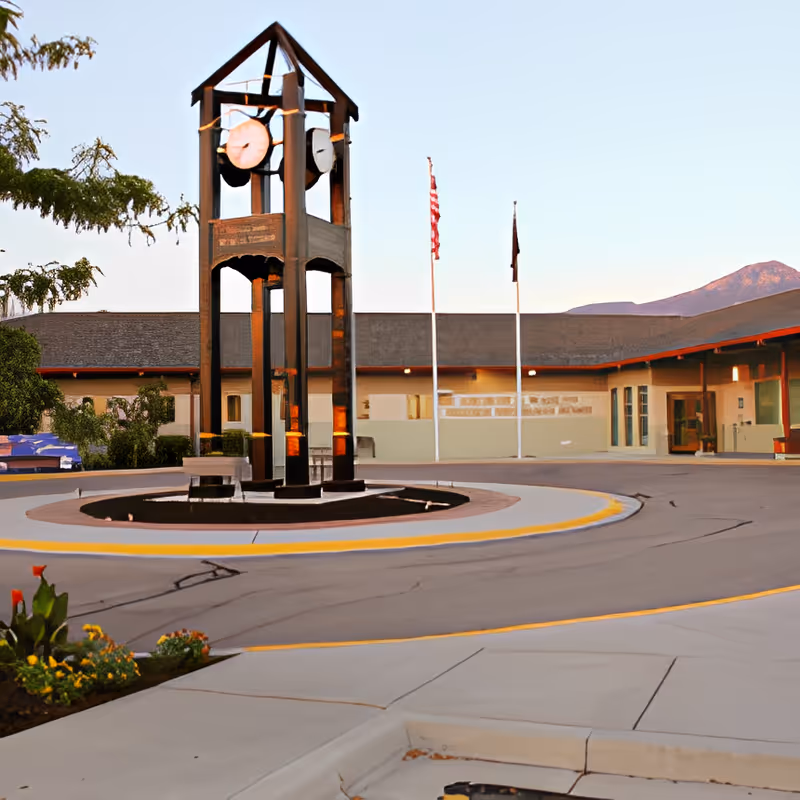 Exterior view of George E. Wahlen Ogden Veterans Home featuring a tall clock tower in the center of a circular driveway, with two flagpoles and a building in the background under a clear sky and mountains in the distance.