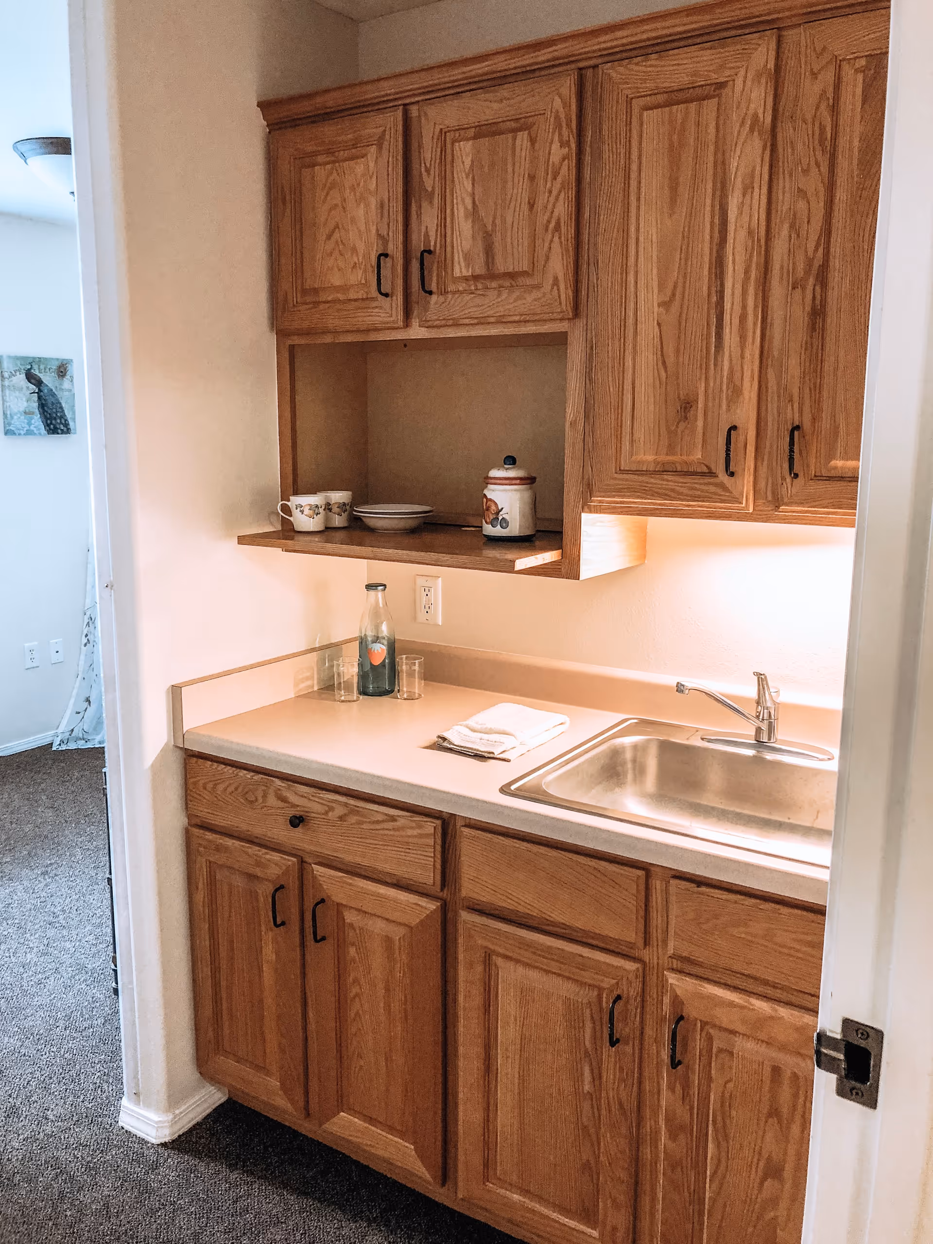 Small kitchen area with wooden cabinets, a countertop with a sink, a glass bottle, two glasses, a folded towel, and some cups and a jar on a shelf. The kitchen opens into a carpeted room with a wall decoration visible.
