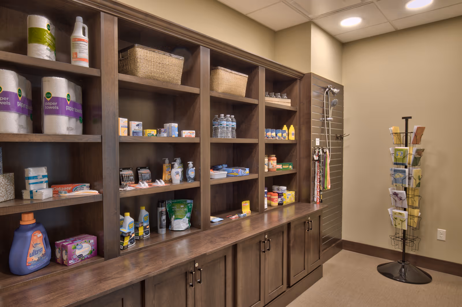 A room with wooden shelves stocked with various household and personal care items including paper towels, cleaning supplies, bottled water, and toiletries. There is a display rack with greeting cards on the right side and a shower head mounted on the wall in the corner.