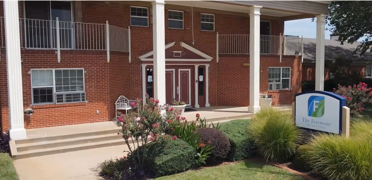 Front exterior view of a two-story brick building with white columns and a porch. There are bushes and flowers in the landscaped area in front of the building. A sign in the grass reads 'The Fairmont Skilled Nursing & Therapy.'