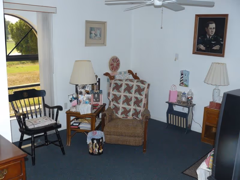 A cozy living room with a brown armchair covered by a quilt, a black wooden chair with a cushion, two side tables with lamps and various decorative items, a ceiling fan, and framed pictures on the white walls. A window with vertical blinds shows a green outdoor view.