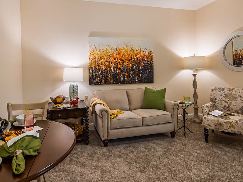 Cozy living room with a beige loveseat flanked by lamps, a patterned armchair, round dining table in the foreground and colorful artwork above the sofa.