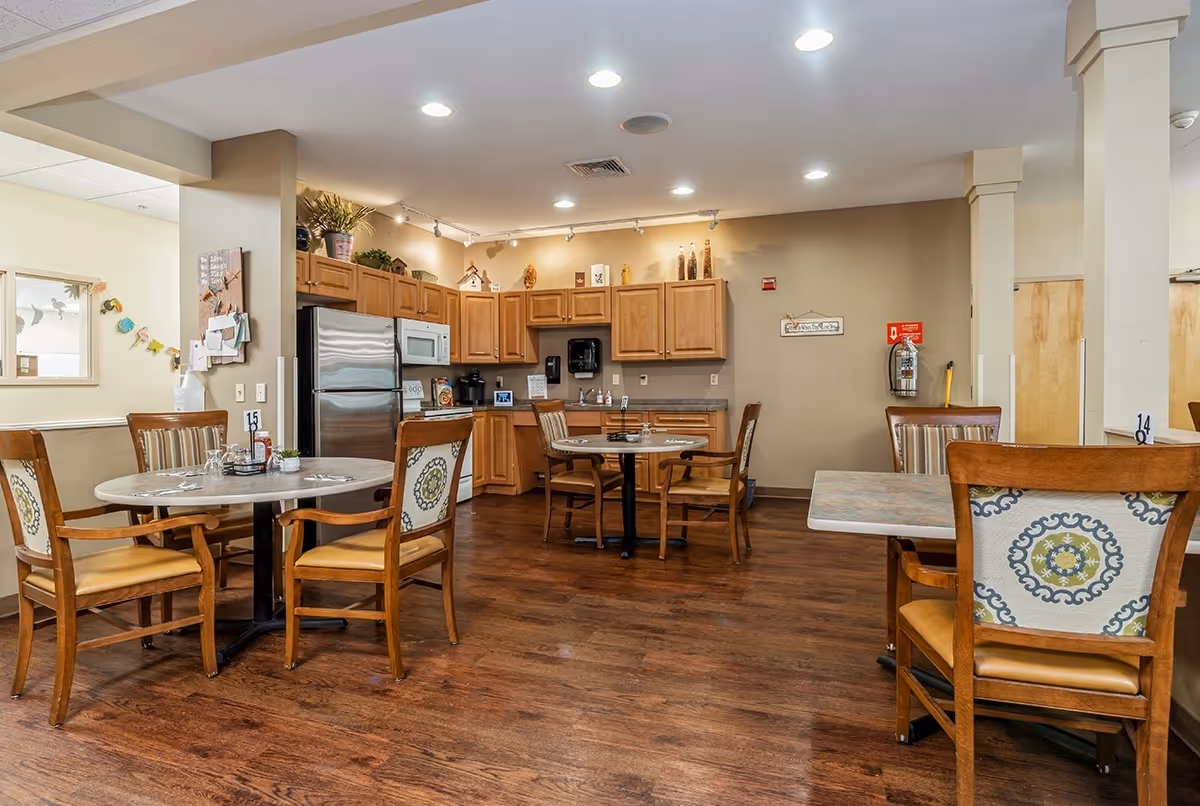 A dining area in a senior living facility with several round and square tables surrounded by wooden chairs with patterned cushions. The room features a kitchen area with wooden cabinets, a stainless steel refrigerator, microwave, and coffee maker. The floor is wooden, and the walls are painted beige with some decorations and a fire extinguisher mounted on one wall.