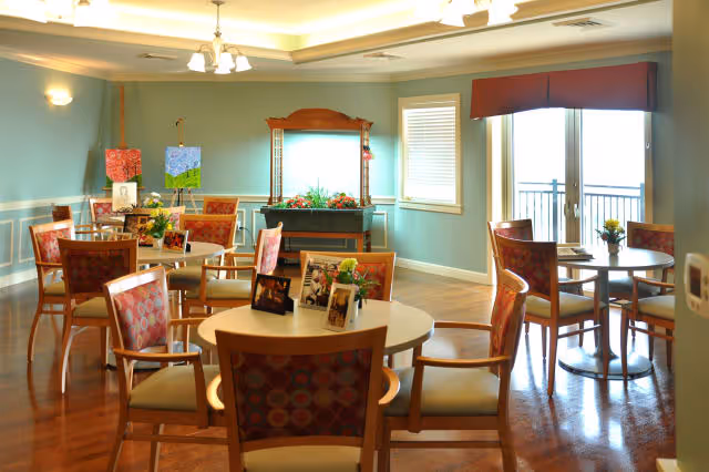 A bright dining room with several round tables and wooden chairs with patterned upholstery. The room has light blue walls with white trim, a large mirror with a wooden frame, and windows letting in natural light. There are small flower arrangements and framed photos on the tables, and a chandelier hanging from the ceiling.