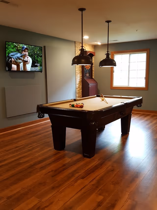 A recreational room with a pool table in the center, two hanging pendant lights above it, a window with wooden trim letting in natural light, a wall-mounted TV showing a man holding a large fish, and a dartboard cabinet in the corner. The room has wooden flooring and gray walls.