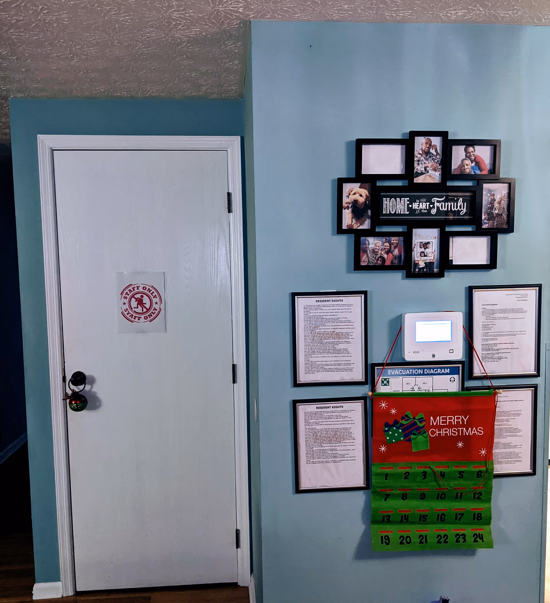 Interior wall of a senior care facility painted light blue with a white door labeled 'Staff Only'. The wall features a collage photo frame with family pictures and the phrase 'Home is the Heart of the Family'. Below the frame are framed documents including resident rights and an evacuation diagram. A red and green Merry Christmas countdown calendar hangs on the wall.