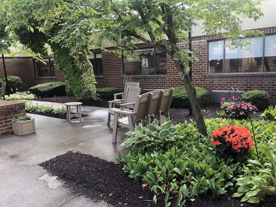 Outdoor garden area at Pulaski Health & Rehabilitation Center featuring a concrete pathway, several wooden chairs and a small table, surrounded by green plants, bushes, and trees with a brick building in the background.